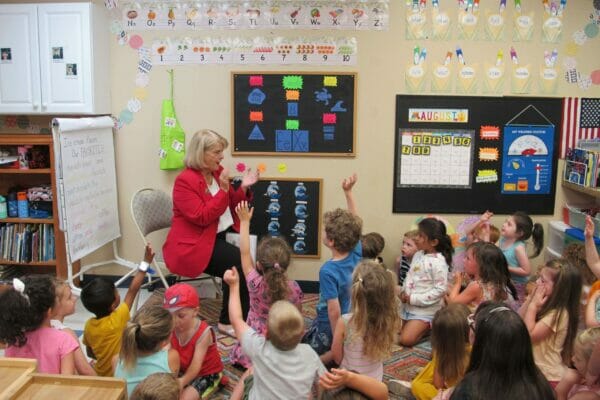 Woman in red jacket answers questions from large group of children with their hands raised.