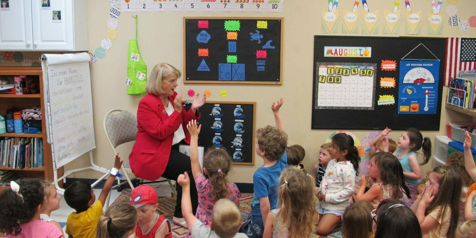 Woman in red jacket answers questions from large group of children with their hands raised.